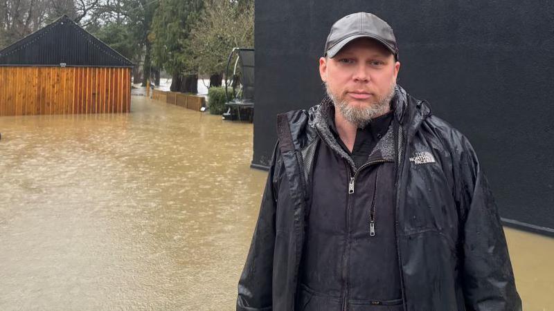 David Graham yn sefyll yn gwisgo ei ddillad gwlyb mewn dwr y tu allan o adeilad.