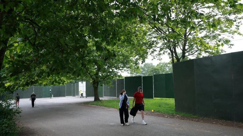 People walk on the road next to Heaton Park. Large black panels of fencing have been put up around the park.