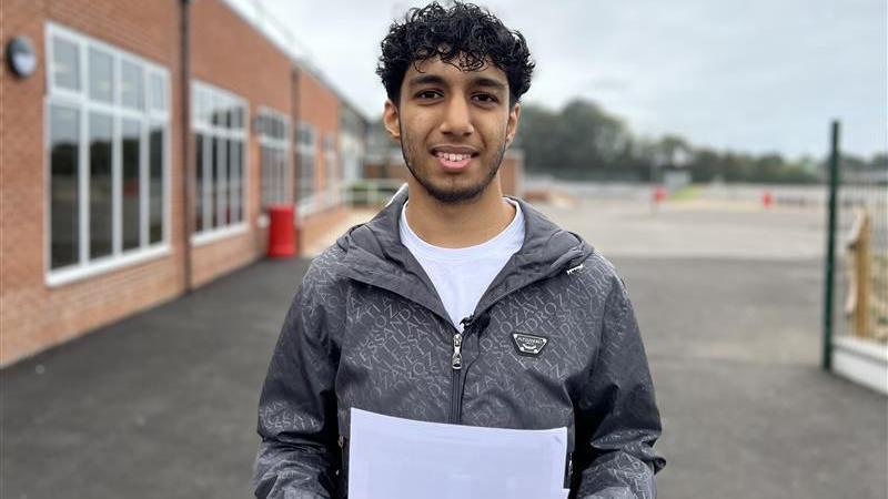 Amaan is standing outside in the school grounds, looking at the camera and holding his results page. He has short black hair, styled into curls on the top of his head. He is wearing a white round neck t shirt with a grey zipped up waterproof jacket. Behind him is a school building to the left and an empty car park.