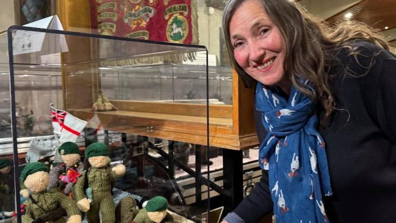 Tansy Forster wearing a blue scarf and navy jumper, pictured beside knitted British soldiers depicting the D-Day landings in Normandy, France, in 1944. The soldiers have been knitted wearing light green uniforms and dark green berets, while their faces and hands are white. The knitted soldiers are also carrying a Union Jack/St George's Cross flag.