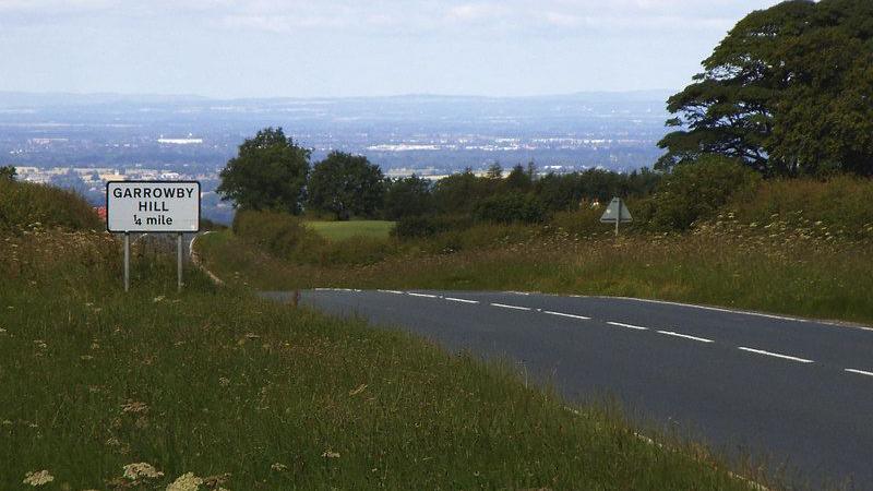 A view of the A166 from the top of Garrowby Hull in East Yorkshire. The road is surrounded by grass verges and fields and trees are visible in the background.