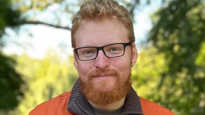 A man with red hair, a beard and black-framed glasses is standing outside and smiling at the camera against a blurred background of trees. 