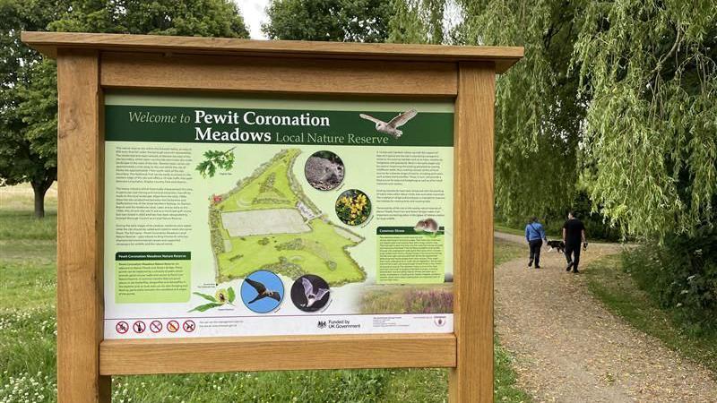 A sign at one of the entrances to the site with a family walking nearby