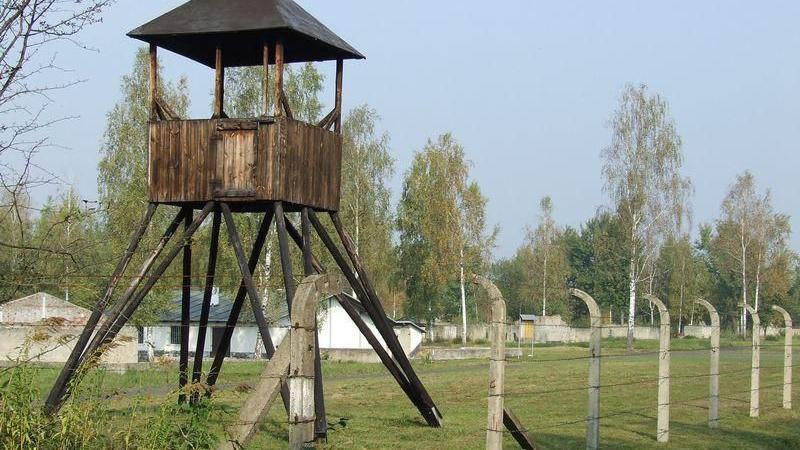 A wooden watchtower in a field behind barbed wire fencing.