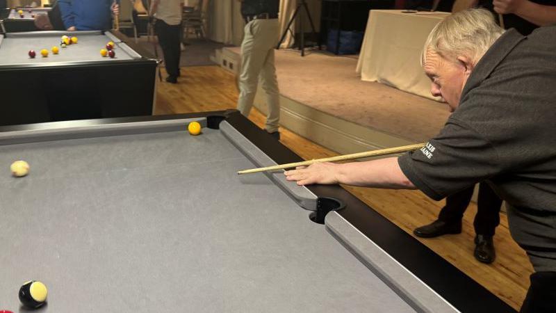 A pool table with a man holding a pool cue taking a shot. He has blond hair and is wearing a dark, short sleeved shirt. The pool table is grey. There is another pool table in the background with people standing around it.