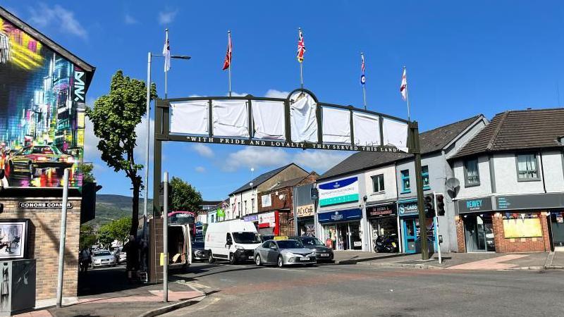 A black arch over a road. There are seven sections in the middle of the top of the arch that are covered in a white cloth. There are five flags along the top.