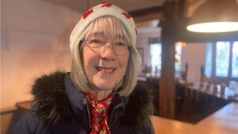 Rachel Radford is looking at the camera. She has shoulder-length fair hair and is wearing glasses, a woolly hat, red scarp and a dark jacket. She is stood in a pub, and a bar can be seen behind her. 
