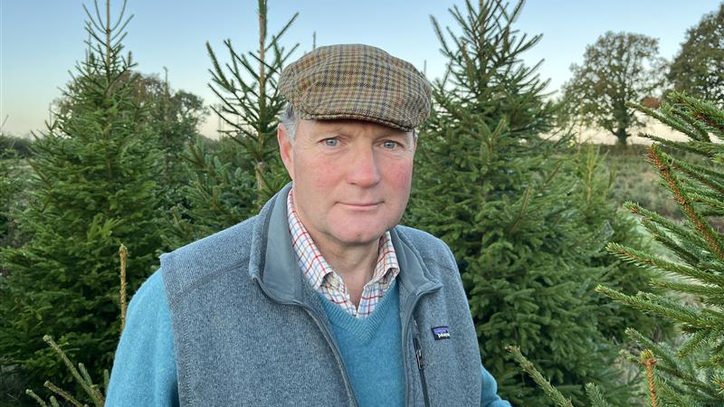 Edward Barnham (a man with a flat cap, a grey fleece, a blue jumper and chequered shirt) stands in front of Christmas trees