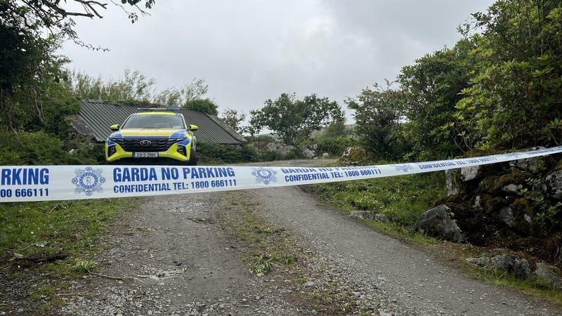 The garda (Irish police) cordon at the scene of a fatal house fire at Gleann Mhic Mhuireann, County Galway. A yellow and blue garda car is parked on a rural country road, lined by trees and shrubs. A corrogated metal roof of a stone building is partially visible behind the car.