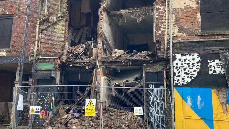 Metal fences sit at the feet of a collapsed building. Fallen brickwork and debris can be seen behind.