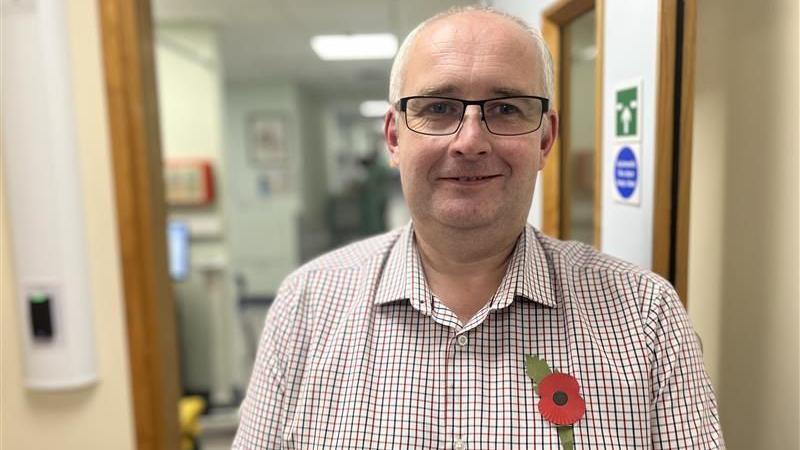 Darryn Allcorn is smiling at the camera. He is standing in the doorway of a hospital room. He is wearing a checked shirt with a remembrance poppy pinned to it and glasses.