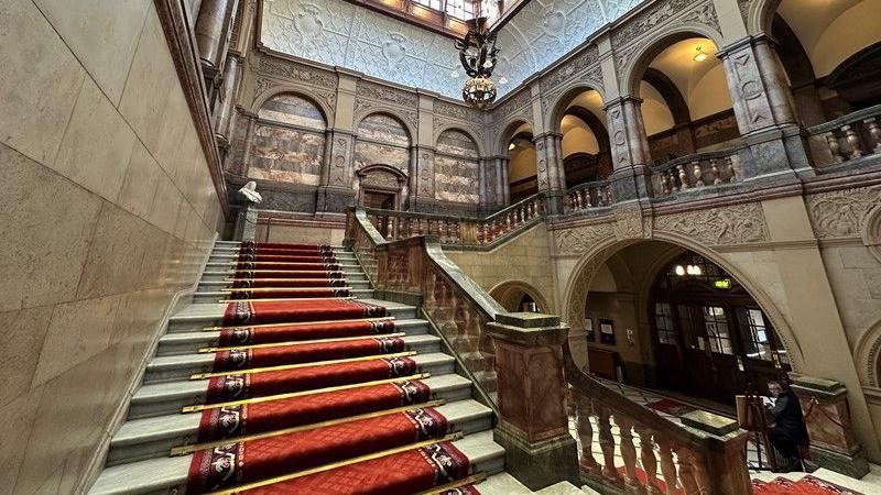 A grand, sweeping marble staircase leads from the front entrance of the town hall upstairs to the council chamber. It has a red carpet running down the middle, with marble banisters. There are ornate marble arches in the town hall foyer and tall ceilings.