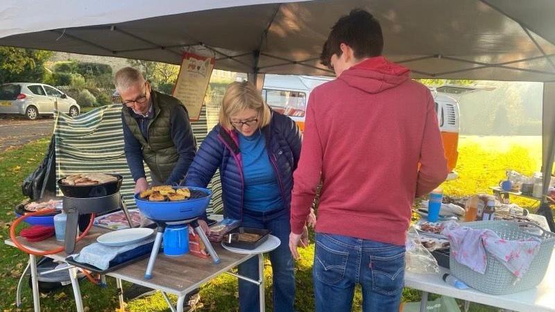 Charlie with mum Rachel and dad Richard making breakfast under a gazebo. His parents are cooking sausages and other food items on two barbeques during a sunny morning. 