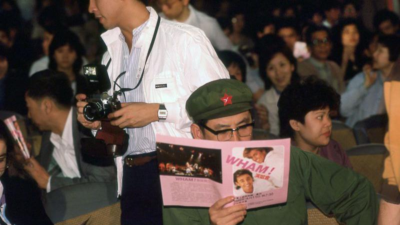 The audience of a Wham! performance in China. One man, dressed in a green uniform, is reading a pink tour programme.