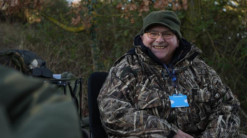 Brian Haycock sitting down and smiling at the camera. He is wearing a camouflage style jacket and a green hat.