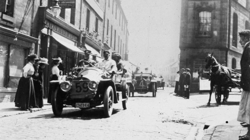 A black and white image of Narrowgate in Alnwick in 1910 with racing cars going through. A number of people are watching from the sides of the road. 