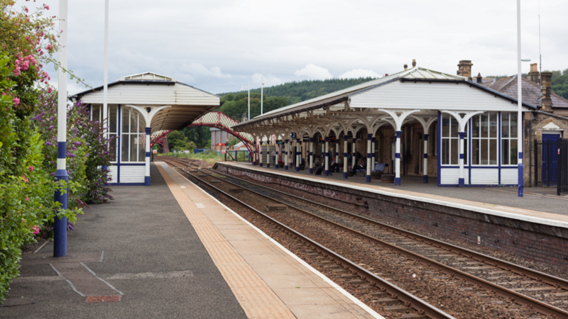 Hexham station with blue and white Victorian station buildings on either side and a red and white footbridge in the distance.