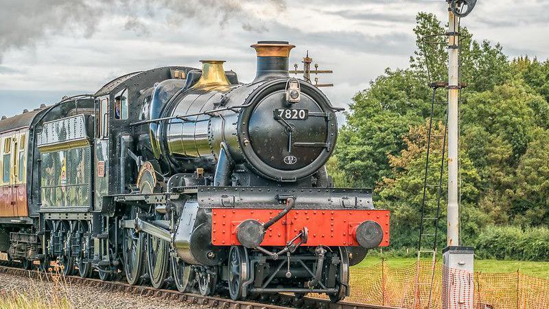 A steam train with a circular face and steam spout and a red bumper on the West Somerset Railway. It's pulling a green coal car, and behind it is a red carriage. 