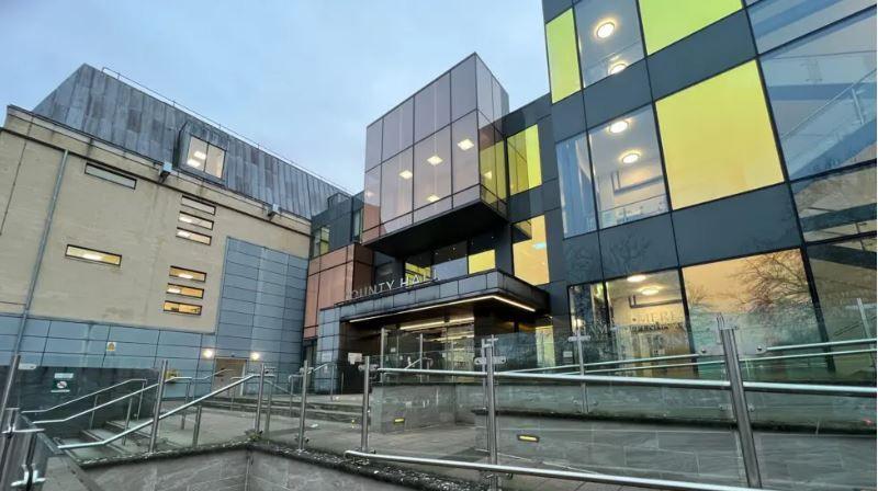 The exterior of County Hall, Trowbridge. The building has a modern facade with large square multicoloured panels in grey, orange and yellow. There are stairs leading up to the front of the building and the words county hall above the entrance.