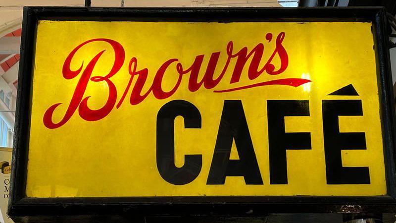 Brown's café sign. The background is yellow with a black outline. The word Brown's is written in red, cursive-style, and the word café is written in black in blocky characters. Liz Woolley