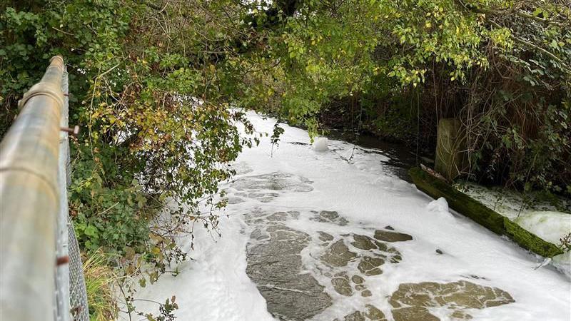 White foam on a river surface. The photo is taken from a bridge and is looking down and along part of the river. Bushes and vegetation line the river, which has a width of just a few metres. 