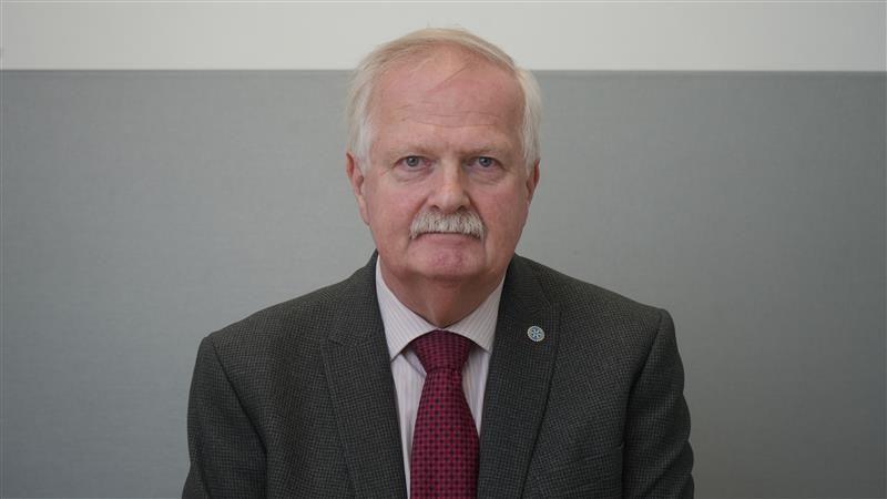 Robin Pivett wearing a suit, shirt and red tie. He has grey hair and a moustache and is looking directly at the camera with a serious expression.