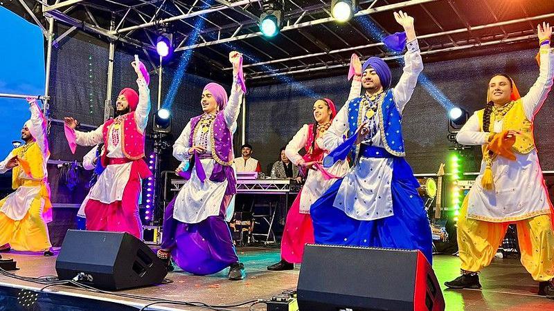 Six bhangra dancers wearing bright orange, pink, purple, blue and yellow traditional clothing, which consists of tops, baggy pants and turbans or stoles over the heads,  perform onstage at the Diwali Mela in Phoenix Park in 2023.