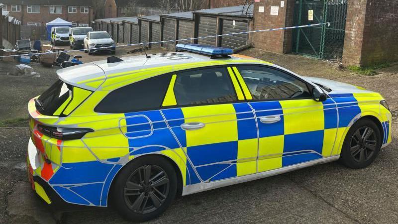 A police car, by police tape, by a row of garages and at the end of the row a police tent and three more vehicles. A pile of rubbish is outside a garage and there are houses at the end of the road. 