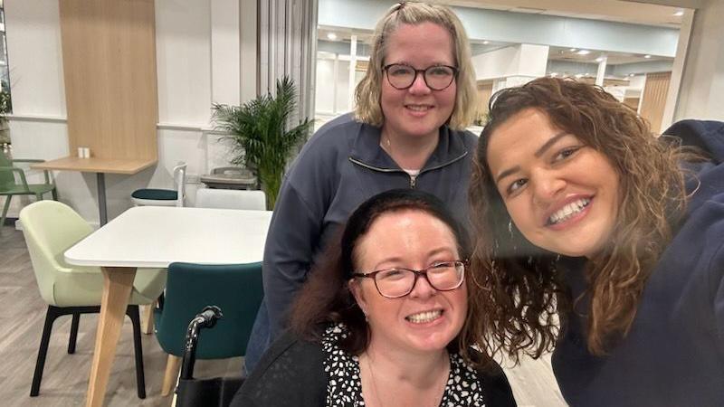 Three woman take a selfie in a cafe. One woman with blonde hair and glasses stands behind another woman with dark hair and glasses who is seated. Another woman with dark curly hair stands and leans in towards both of the other women.