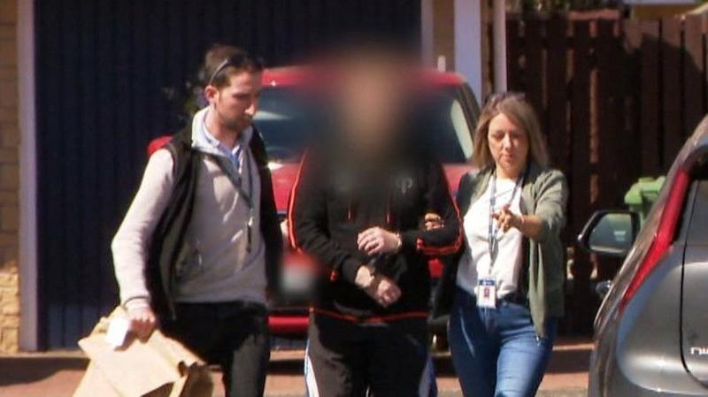 Two plain clothed detectives lead away a man into an unmarked police car in a suburban housing estate. The officers - one male, one female are in jeans and trainers. The suspect is in trainers and tracksuit bottoms and has been blurred to hide his identity.