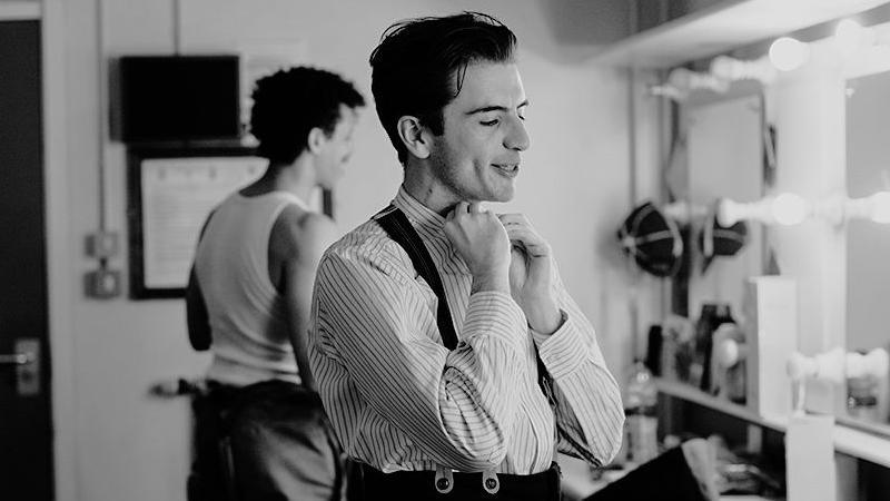 Rhys Warrington, wearing a striped shirt and braces, seems to be buttoning the collar of his shirt. He is stood in a dressing room, with a mirrored dressing table to his right and another man stood nearby is looking towards the mirror.