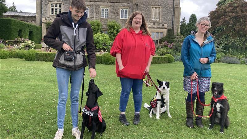 Three trainers stand in a line smiling with their dogs, who are all sitting by their sides 