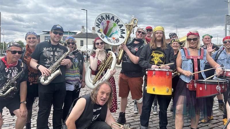 A group of people - all in a band - stand on a cobbled street smiling at the camera holding instruments including drums and trumpets. The sky is cloudy. 