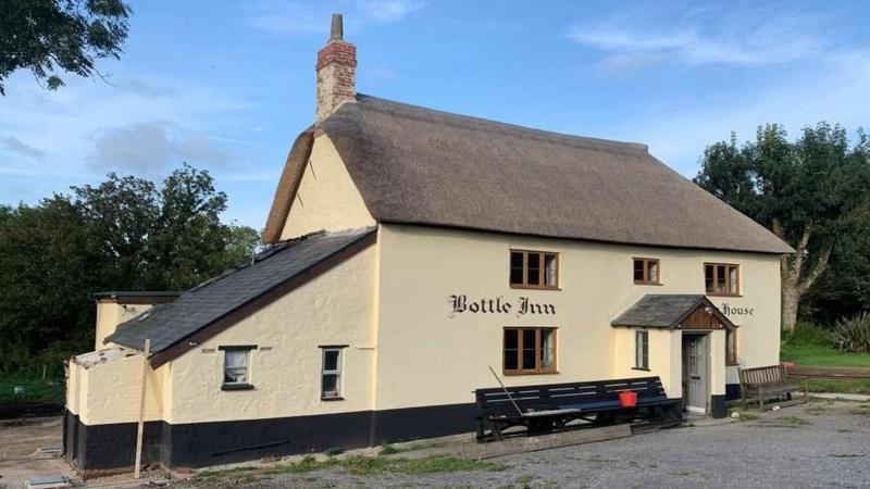 The newly painted and newly thatched Bottle Inn pub. The pub is a double-fronted detached building, painted cream, with a small porch, a thatched roof and a lean-to structure on the left side.