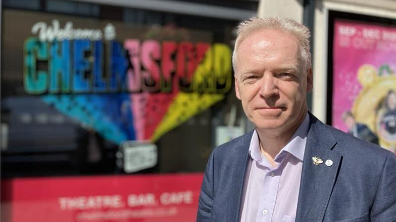 Stephen Robinson is in a navy blue suit jacket and a purple shirt, standing outside, with some colourful Chelmsford branding behind him.