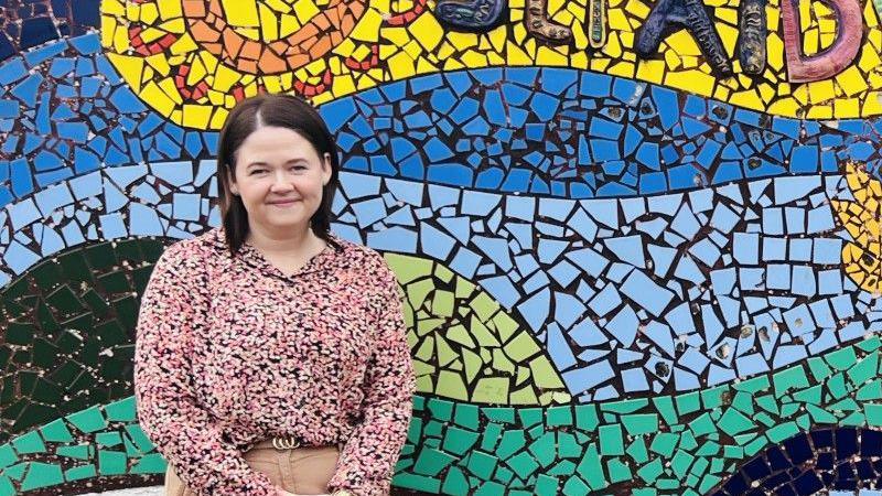 Judith is wearing a pink flowery shirt and beige chinos. She has a gold belt on. Her hair is brown and to her shoulders and she is smiling at the camera. She is standing in front of a blue, yellow and green background. 