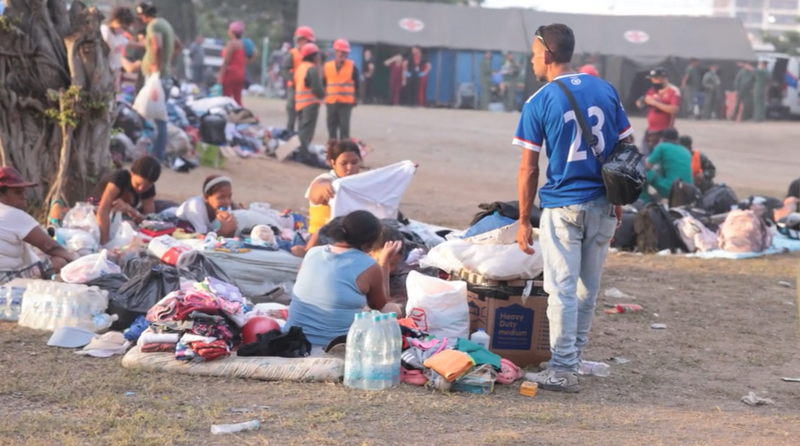 People sit on the ground, surrounded by their possessions, with an emergency tent in the background.