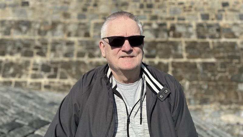 Man with short grey hair smiling at the camera with sunglasses on. He is wearing a black coat and a grey zip up jumper underneath.