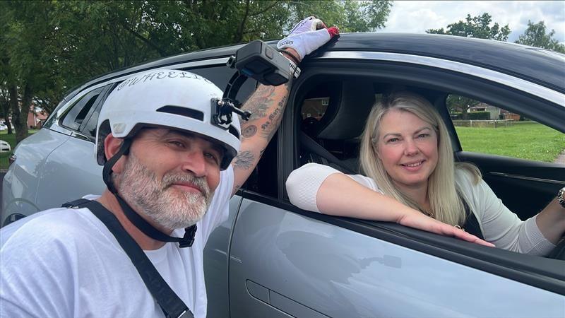 Mr Parker sits in his wheelchair next to a chair that his partner Louise sits in. He rests an arm on the top of the car. Louise sits in the driver's seat and rests her elbow on the open window. She has long blonde hair and wears a white long-sleeved top.