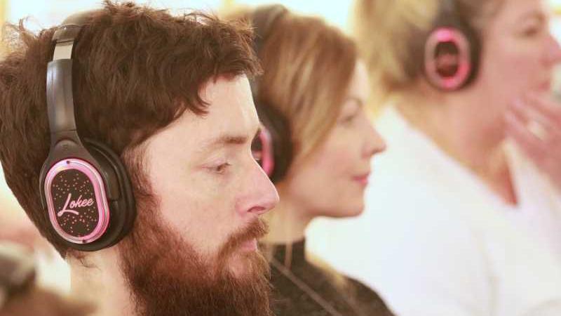 Alex Innes wearing headphones that are black and pink. He has a dark beard and dark hair. Two other women are to the right of the picture, but they are in the background and slightly blurred. They all have headphones on.