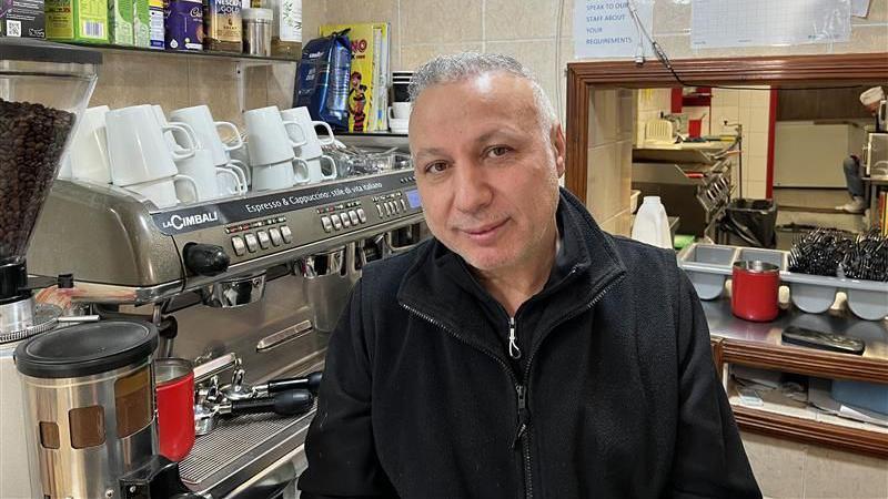 A man looking to camera wearing a black top in a cafe with a drinks machine behind him 