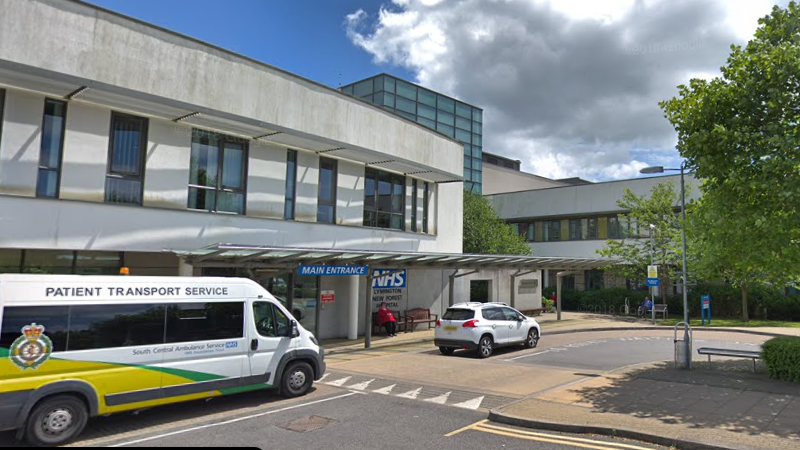 An ambulance and a car are parked outside the main entrance to Lymington New Forest Hospital - a grey, two-storey building with a large blue NHS sign on the front