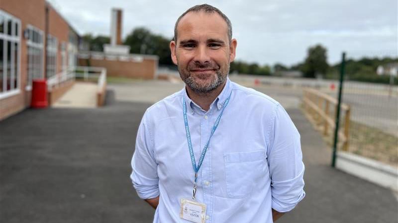 Andy Murdock is standing outside in the school grounds, looking at the camera and smiling, with his arms clasped behind his back. He has very short greying black hair, and some stubble. He is wearing a white collared shirt, rolled up at the sleeves and a pale blue lanyward around his neck. Behind him is a school building to the left and an empty car park.