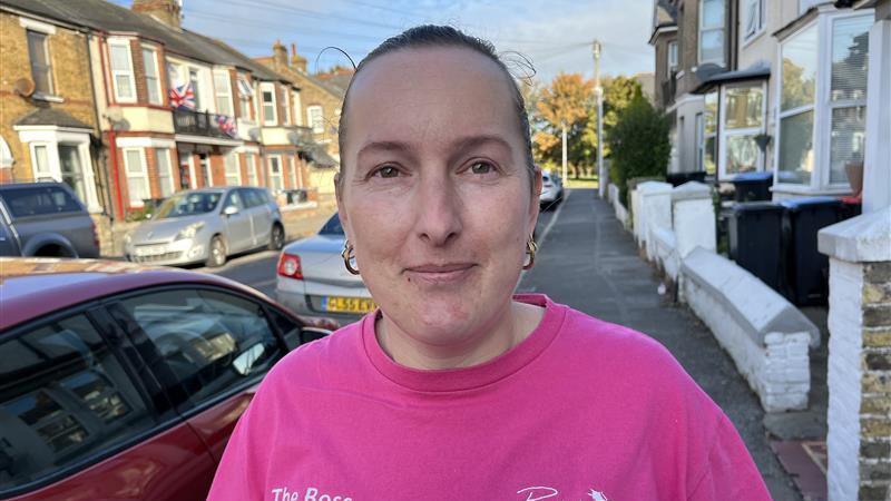 Laura Burbridge wears a bright pink t-shirt as she poses for a picture in a terraced road in Margate. She is a young woman with her light brown hair scraped back.