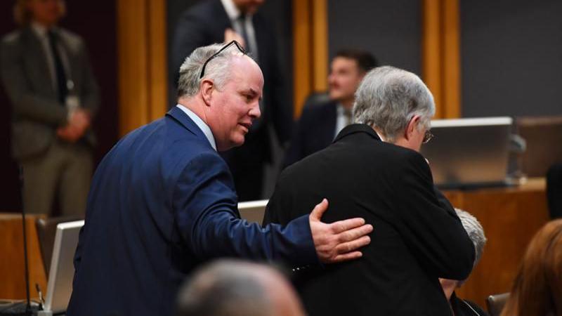 Andrew RT Davies with his hand around Mark Drakeford in the Senedd debating chamber. Davies is wearing a dark blue suit while Drakeford is wearing a black suit. His face is out of shot but Davies's face can be seen side on.