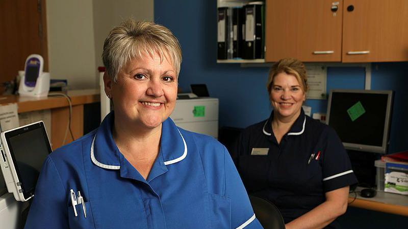 Two women in blue nurses' uniforms sit and smile at the camera. One woman is closer to the camera, she has short grey hair. The woman on the right sits further back and has blonde hair tied back. They sit in a room with a computer, printer, and medical equipment in it.