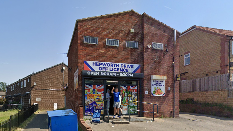 A two story brick building with a blue and white sign reading Hepworth Drive Off Licence above a doorway with large windows either side displaying posters for alcohol and grocery products.
A man dressed in t shirt and shorts stands in the doorway.