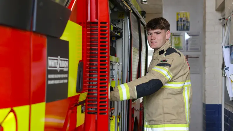 Bobi Jones smiles as he stands beside a fire engine in his firefighter's uniform. 