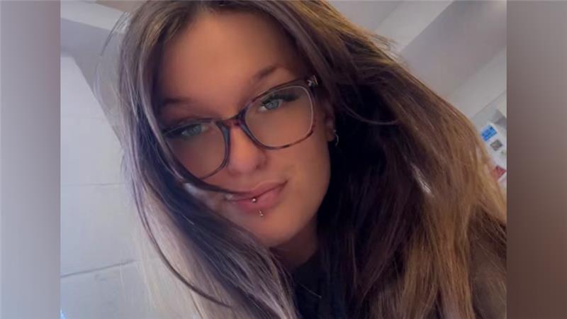 A selfie of a woman with long brown hair and brown glasses. She has a piercing through her bottom lip