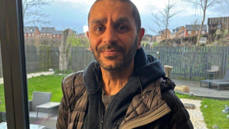 Satpal is standing in the middle of the photo. In the background is a garden. He is wearing a black hoodie and grey T-shirt.